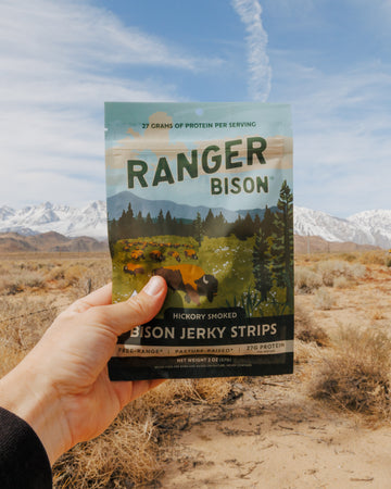 Hand holding a package of Ranger Bison jerky against a scenic background with mountains and sky.